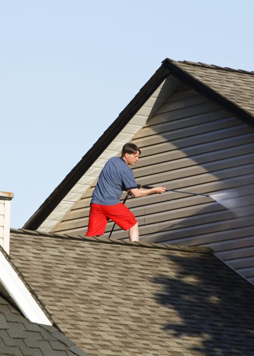 A man standing on the roof of his house cleaning the vinyl siding.