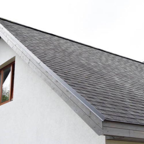 Fragment of white house with grey asphalt shingles roof with just installed wooden eaves and brown window on decorative plaster facade with copy space for text.